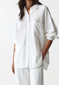 Camicia bianca, morbida e ampia, con bottoni, colletto, una tasca sul petto e maniche arrotolate, realizzata in tessuto leggero e dalla texture evidente.