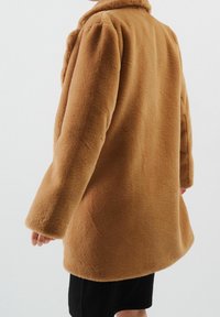 Abrigo marrón de textura suave con un corte relajado, que presenta un cuello y mangas anchas, terminando por encima de las caderas. Diseño simple y limpio.