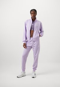 Το λιλά αθλητικό σετ περιλαμβάνει ένα σακάκι με φερμουάρ και εφαρμοστά παντελόνια, φτιαγμένα από ένα λείο υλικό. Διαθέτει το λογότυπο της Adidas σε λευκό χρώμα.