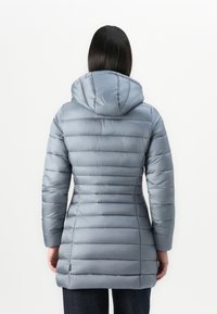 Vrouw in een lange, lichtblauwe pufferjack met capuchon, gezien van achteren tegen een effen witte achtergrond.