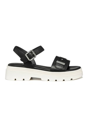Sandalia de cuero negro con hebilla plateada en la correa del tobillo, correa frontal ancha con detalle decorativo y suela gruesa blanca con estrías.