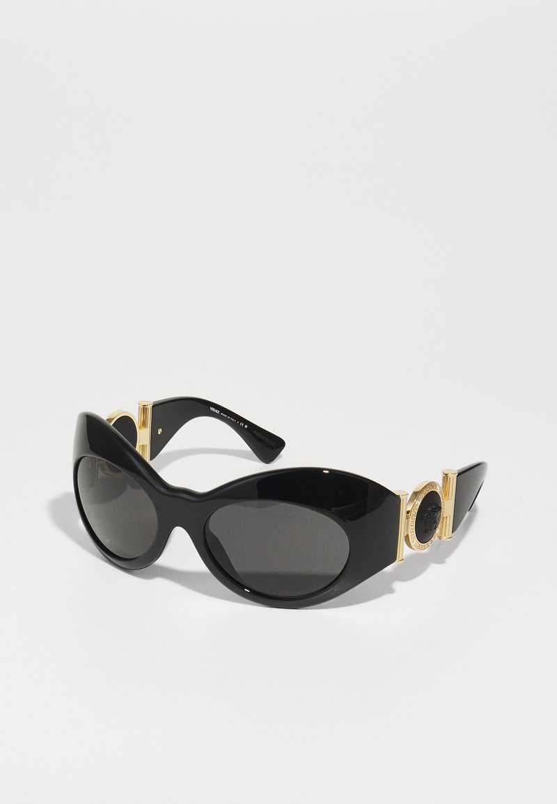 Lunettes de soleil noires avec une finition brillante, de grande taille en forme d'œil de chat, verres foncés et éléments en laiton avec un emblème rond sur les branches.