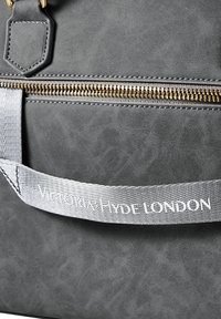 Grå kunstlæder håndtaske med en gylden lynlås, der har en tekstureret overflade. En sølvfarvet rem viser "Victoria Hyde London."