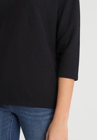 Schwarzes, strukturiertes Oberteil mit Dreiviertelärmeln, kombiniert mit blauen Jeans. Der Stoff hat ein gewebtes Muster, das eine subtile Tiefe und Kontrast bietet.