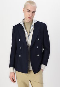 Blazer doppiopetto blu marino con bottoni argentati, abbinato a un maglione crema e una camicia a fantasia floreale, pantaloni khaki e polsini a coste.