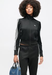 Μαύρο αθλητικό μπουφάν με ψηλό γιακά, κλείσιμο με φερμουάρ και τρεις λευκές ρίγες στα μανίκια. Συ νδυασμένο με μαύρο παντελόνι και μπλε τσάντα.