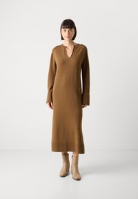 Abito lungo a maniche lunghe in maglia marrone con scollatura a V profonda, polsini a coste e orlo a coste. Indossato con stivaletti alla caviglia beige, caratterizzati da una texture liscia.