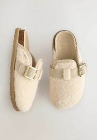 Sabots en tissu beige duveteux avec une sangle et une boucle dorée. Surface texturée avec une semelle plate, adaptés à un usage intérieur. Pas de motifs visibles.