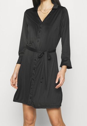 Robe-chemise noire à manches longues avec des boutons sur le devant et une ceinture à la taille, portée avec des colliers dorés superposés.