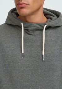 Sweatshirt à capuche gris avec une texture douce, présentant des cordons de tirage crème clair et des œillets en métal, mettant en avant un design décontracté et confortable.