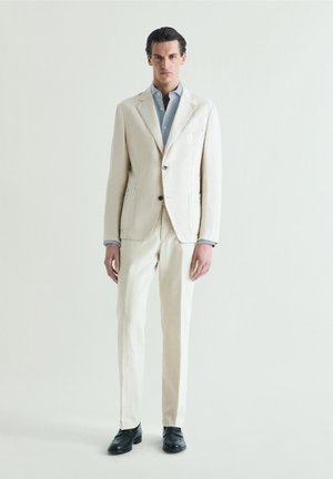 Hombre con un traje color marfil, camisa azul clara y zapatos negros, de pie contra un fondo claro y liso.