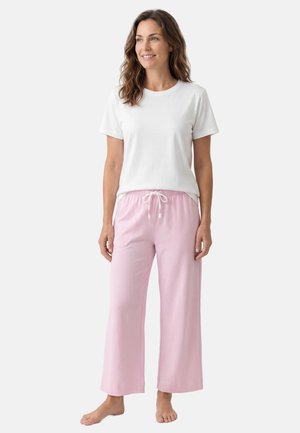 Frau steht barfuß, trägt ein weißes T-Shirt mit kurzen Ärmeln und hellrosa weite Hosen mit Kordelzug vor einem einfarbigen Hintergrund.