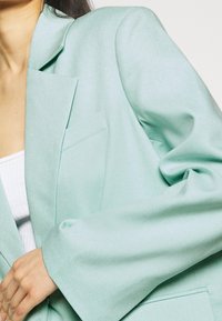 Blazer verde menta con rever a punta, tasca singola e vestibilità ampia. Tessuto liscio e maniche leggermente oversize.