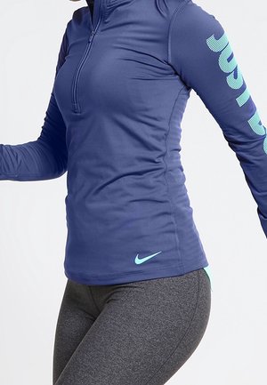 Persoon die een aangesloten blauwe lange mouwen sporttop met een voorrits en grijze legging draagt, in beweging tegen een effen achtergrond.