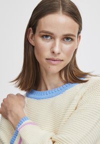 Pullover mit einer cremigen Stricktextur, blauem geripptem Halsausschnitt und Bündchen sowie rosa Akzenten an den Ärmeln. Nahaufnahme der Vorderansicht.