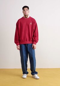 Felpa rossa con colletto a girocollo e patch con logo, abbinata a jeans blu larghi e sneakers bianche. Texture lisce e vestibilità comoda.