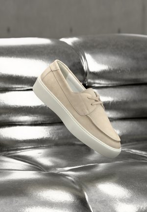 Beige suede slip-on shoe with a white sole, featuring a rounded toe and minimalistic design, set against a shiny metallic background.