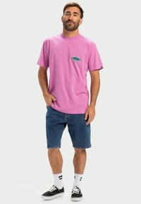 Roze katoenen T-shirt met korte mouwen en een blauwe diamantgrafiek op de borst, gecombineerd met denim shorts en zwarte sneakers.