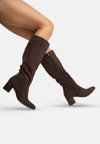 Botas altas de ante marrón hasta la rodilla con tacón cuadrado y cremallera lateral; diseño ajustado con una textura suave y punta afilada.