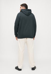Felpa con cappuccio in tessuto verde scuro, con maniche lunghe e vestibilità comoda. Abbinata a pantaloni color crema e scarpe nere.