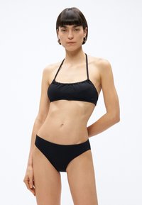 Kobieta o krótkich brązowych włosach, ubrana w czarny, fakturowany bikini z wiązanym na szyi topem i dopasowanymi majtkami, stojąca na białym tle.