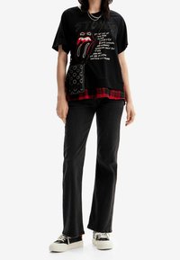 Camiseta gráfica negra con el logo rojo de los Rolling Stones, superpuesta sobre una camisa de cuadros roja. Combinada con jeans negros acampanados y zapatillas de plataforma blanca.