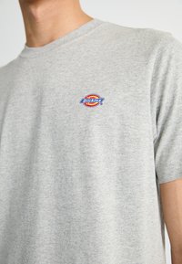 Pilkas medvilnės marškinėliai su apvali apykakle, trumpomis rankovėmis, su spalvingu Dickies logotipu kairiajame krūtinės šone. Sumatryta tekstūra.