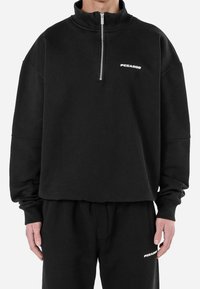 Felpa nera con zip e colletto alto, realizzata in materiale morbido. Presenta un logo bianco sul petto e polsini a costine per una vestibilità aderente.