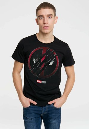 Young man wearing black Marvel Studios Deadpool t-shirt with claw marks design, standing with hands in blue jeans pockets.