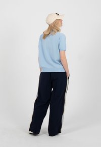 Personne aux longs cheveux blonds portant une casquette beige, une chemise bleu clair à manches courtes et un pantalon noir avec des bandes latérales, debout de dos.