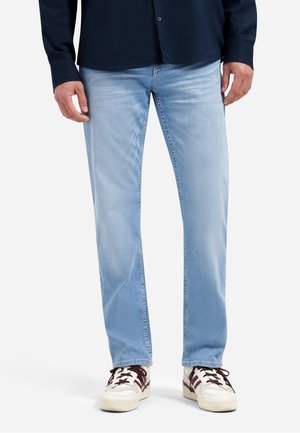 Man wearing light blue straight-leg jeans, white sneakers with dark stripes, and a dark long-sleeve shirt, standing against white background.