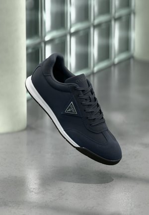 Dunkelblauer Sneaker mit weißer Sohle und schwarzer Laufsohle, mit Guess-Logo an der Seite, vor einem Hintergrund mit gefliester Glaswand dargestellt.
