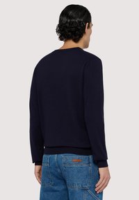 Marineblauer Pullover mit langen Ärmeln, gerippten Bündchen und Saum, glatter Textur. Über blauen Jeans getragen, die mit aufgesetzten Taschen und Ziernähten versehen sind.