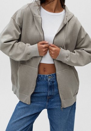 Persoon draagt een grijze hoodie met rits over een wit crop top en blauwe high-waisted jeans, de handen houden de rits van de hoodie dicht bij de borst vast.