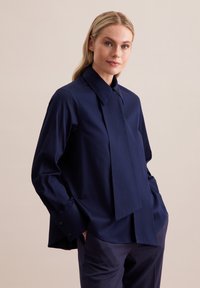 Marineblauwe blouse met lange mouwen, brede kraag, voorplooien en knoopmanchetten. Soepele stof met een relaxte pasvorm. Gecombineerd met bijpassende broek.