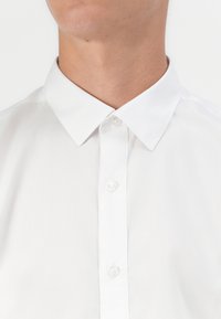 Gros plan d'une personne portant une chemise blanche impeccable à col avec les boutons fermés, montrant le cou et la partie supérieure de la poitrine.