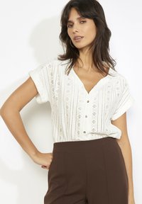 Balta blūze ar V veida izgriezumu, īsām uzlocītām piedurknēm un smalkiem caurumu rakstiem. Kombinēta ar augstām brūnām biksēm.