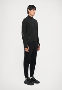Svart sammenleggbar jakke med høy krage, frontlukking med glidelås og refleksdetaljer, kombinert med svarte joggebukser og svarte joggesko.
