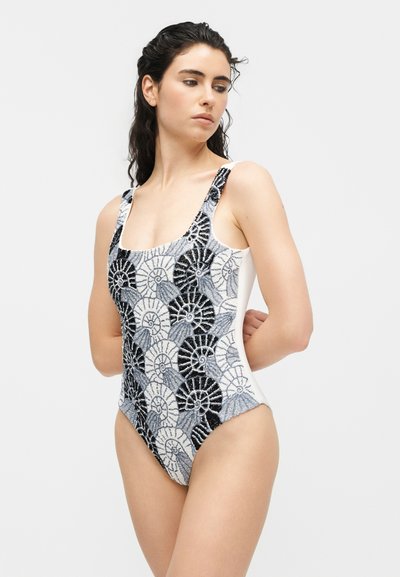 Γυναίκα με μαύρο, λευκό και γκρι μαγιό χωρίς μανίκια με μοτίβο που μοιάζει με κέλυφος, στέκεται με τα χέρια πίσω από την πλάτη, κοιτάζοντας προς τα πλάγια σε απλό φόντο.