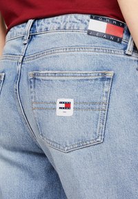 Jeans de denim azul claro con costuras visibles, presentando una etiqueta blanca en el bolsillo trasero con elementos de diseño en rojo y azul y marca.
