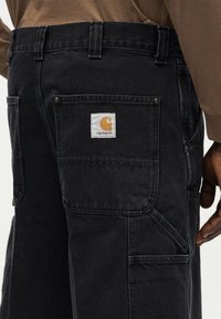 Μαύρο παντελόνι εργασίας Carhartt με πολλές τσέπες και ορατό σημάδι λογότυπου στην πίσω τσέπη, φορεμένο με καφέ πουκάμισο μακρύ μανίκι.