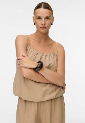 Femme aux cheveux coiffés en arrière, portant un haut beige sans manches et un pantalon, un bracelet noir et des boucles d'oreilles noires, bras croisés, regardant la caméra.