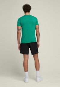 Camiseta atlética verde de manga corta hecha de una tela suave, combinada con pantalones cortos negros. También son visibles zapatillas blancas y calcetas blancas acanaladas.