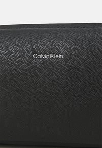 Sac en cuir noir avec une surface texturée et un logo embossé "Calvin Klein" en argent. Forme rectangulaire avec un design épuré.