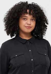 Black button-up shirt with a collar, two chest pockets, and snap-button closures. Made of smooth fabric, features a relaxed fit and texture.