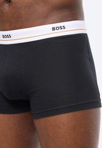 Fekete, testhezálló boxeralsó fehér derékpánttal, amelyen a "BOSS" márkanév olvasható, és egy sötét bőrű személy viseli.