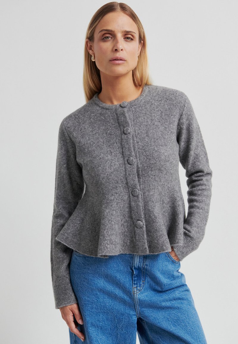 Cardigan gris en maille avec encolure ronde, cinq boutons sur le devant et ourlet évasé en bas, associé à un jean en denim bleu.