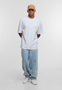 Gray short-sleeve t-shirt, light blue baggy jeans, white sneakers, beige cap, and clear glasses, standing against a neutral background.