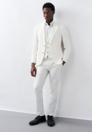 Άνδρας που φοράει λευκό κοστούμι τριών τεμαχίων και μαύρα παπούτσια, στέκεται με το ένα χέρι στην τσέπη, μπροστά σε έναν λευκό τοίχο.