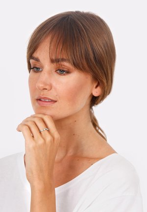 Mlada žena svijetlosmeđe kose i plavih očiju, odjevena u bijelu majicu, naslanja bradu na ruku pokazujući srebrni prsten na prstu.